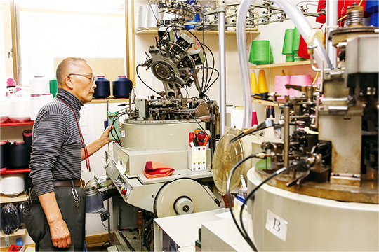この道50年以上の職人・新堂さん。丸編み機の具合を見守りながら、糸の調整などを行います